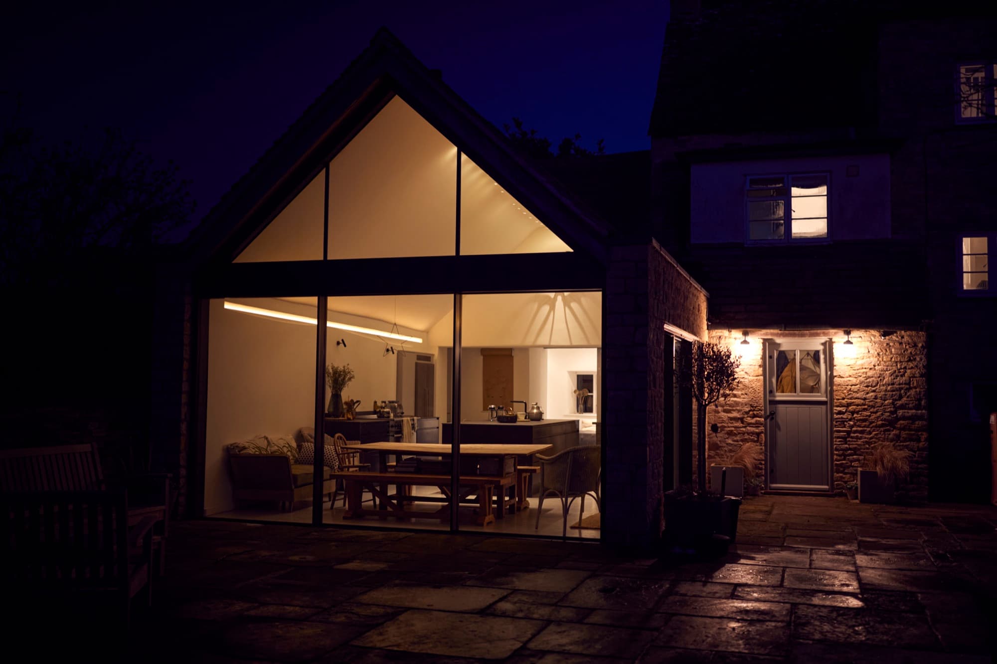 Gabled glazed kitchen extension lit from within at dusk, seen across a stone patio from the garden